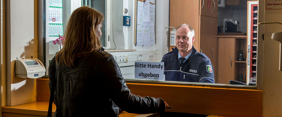 Eine Besucherin einer JVA gibt an der Besucherpforte ihr Mobiltelefon an den Pfortenbediensteten ab.